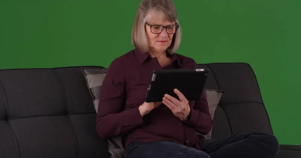 Modern elderly female sitting on couch using tablet device on green screen