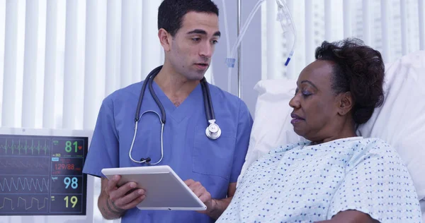 Senior female black patient lying in hospital bed listening to doctor talk