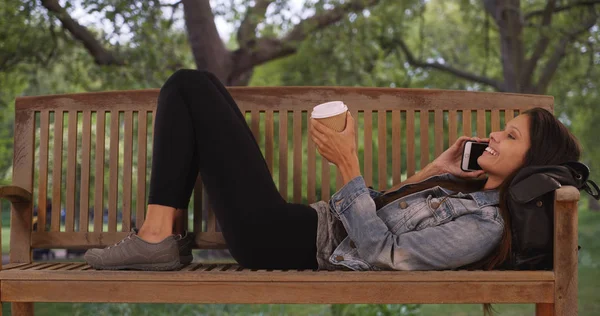 Millennial woman lying on park bench under trees while talking on cell phone