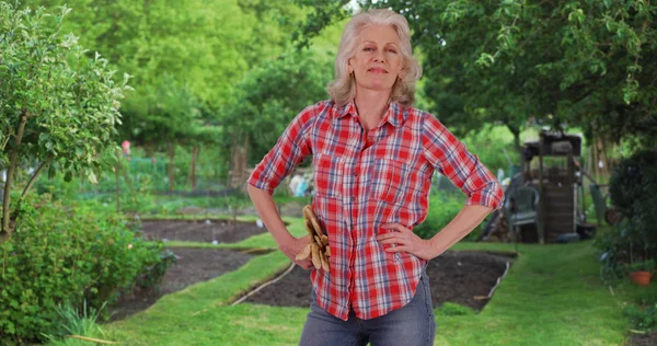 Mature woman in red flannel shirt holding gloves smiles at camera in garden