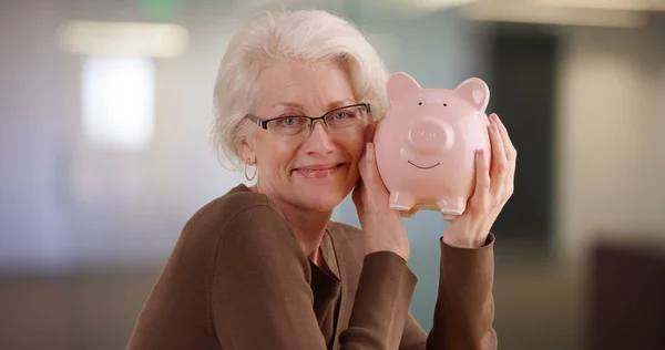 Caucasian elder woman holding piggy bank posing happily for camera indoors