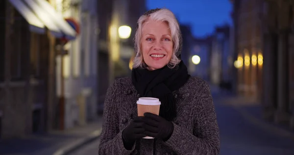 Happy older woman in sweater and scarf holding paper coffee cup at night