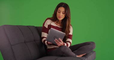Portrait of pretty Latina using tablet sitting on couch on green screen clipart