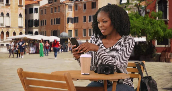 ヴェネツィアのテキスト メッセージの携帯電話上での街のカフェのテーブルに座っているかわいい黒人女性 — ストック写真