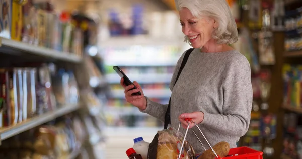 Happy senior woman looking at grocery store aisle with cellphone in hand