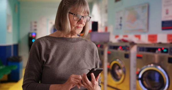 Somber old lady reading distressing news alone at laundromat