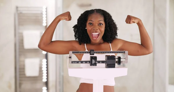 微笑的黑人女子站在体重秤上欢呼 炫耀强壮的手臂 — 图库照片