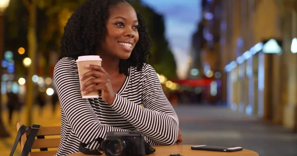 Smiling black woman sits at cafe table on Champs Elysees street looking around
