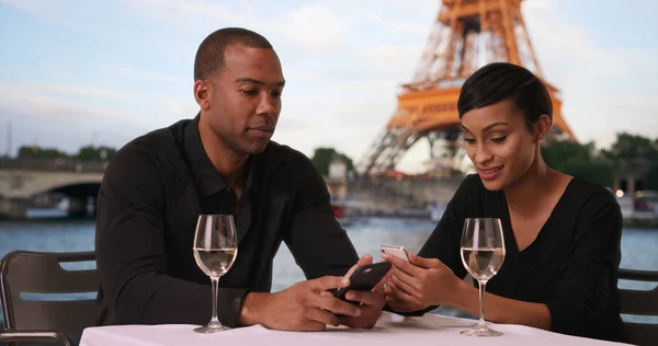 Dating African couple using phones while sitting outside near Eiffel Tower