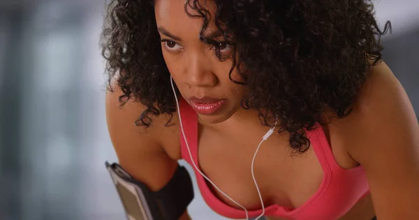 Strong African-American fitness girl with earphones in breathing heavily indoors