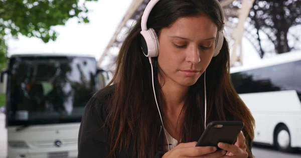 Millennial tourist woman listens to music and texts by tour bus depot in Paris