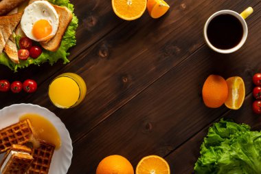 Kızarmış yumurtalı tost, sosis, taze marul ve vişneli domates. Kahve, cam bardakta meyve suyu, waffle ve ahşap arka planda portakal dilimleri. Amerikan kahvaltısı, üst manzara, kopyalama alanı