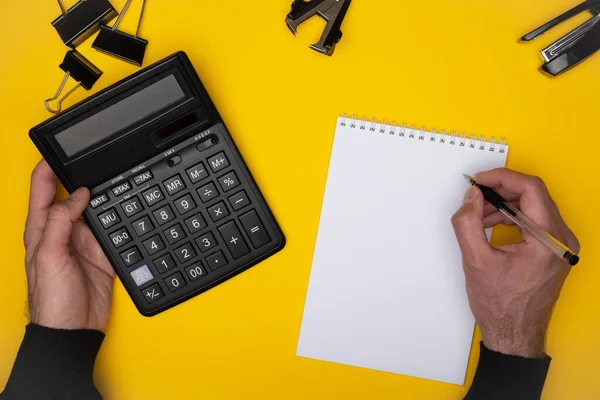 Top view of black calculator and pen in man sleeve up hands writing in notepad with stationery clip, stapler, staple remover on yellow background. Business, finance, stationary, education concept.