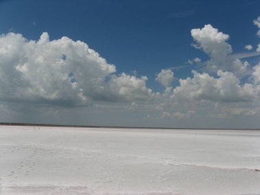 Ufkun ötesine uzanan kurumuş bir tuz gölü. Mavi gökyüzü ve bulutların arka planında beyaz tuz. Beyaz, tuzlu çöl. Mavi gökyüzü ve beyaz bulutlar. Tuz gölünün arka planındaki ufuk çizgisi.