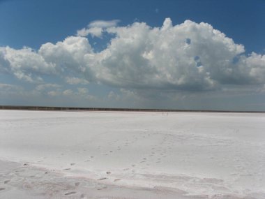 Ufkun ötesine uzanan kurumuş bir tuz gölü. Mavi gökyüzü ve bulutların arka planında beyaz tuz. Beyaz, tuzlu çöl. Mavi gökyüzü ve beyaz bulutlar. Uzaklara giden insan izleri..