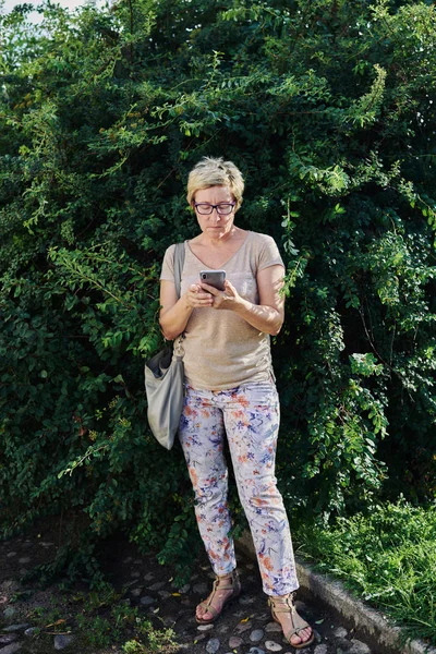Старша жінка використовує смартфон біля куща — стокове фото