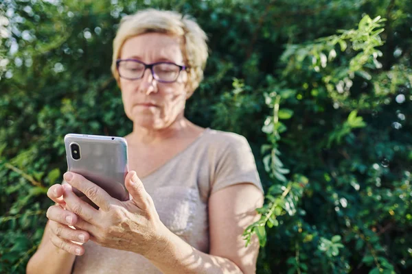 Mujer mayor usando smartphone cerca de arbusto — Foto de Stock