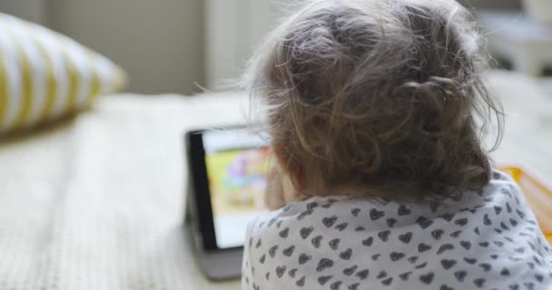 Kind liegt auf dem Bauch auf dem Bett und schaut Zeichentrickfilme auf dem Tablet — Stockvideo