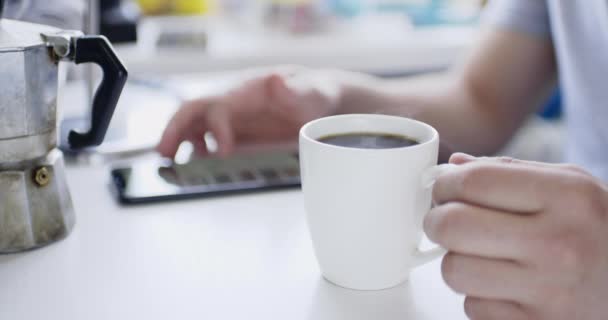 El hombre se sienta a la mesa con el teléfono inteligente y bebe bebida caliente fresca — Vídeos de Stock