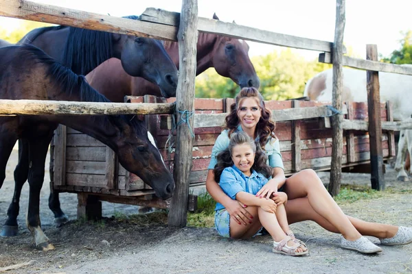 Маленькая улыбающаяся девочка с мамой с вьющимися волосами, одетыми в джинсы, сидящая на земле возле забора у конюшни — стоковое фото