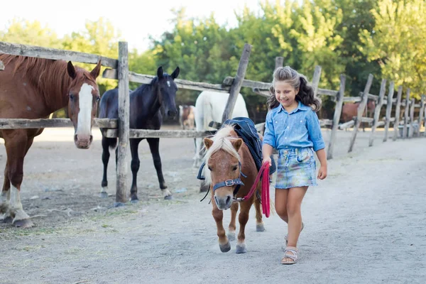 Une petite fille souriante aux cheveux bouclés habillée en jeans marchant avec un poney à l'écurie — Photo