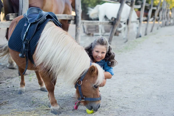 Маленькая улыбающаяся девочка с кудрявыми волосами, одетая в джинсы, гуляющая с пони в конюшне — стоковое фото