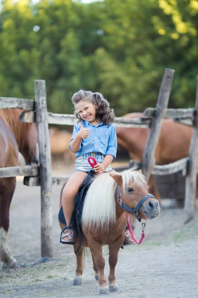 Uma pequena menina sorridente com cabelo encaracolado vestida de jeans montando um pônei no estábulo — Fotografia de Stock