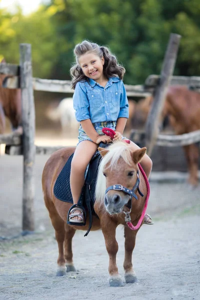 Una pequeña chica sonriente con el pelo rizado vestida con vaqueros montando un pony en el establo — Foto de Stock