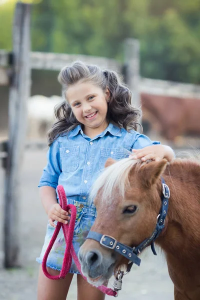 Une petite fille souriante aux cheveux bouclés habillée en jeans avec un poney à l'écurie — Photo