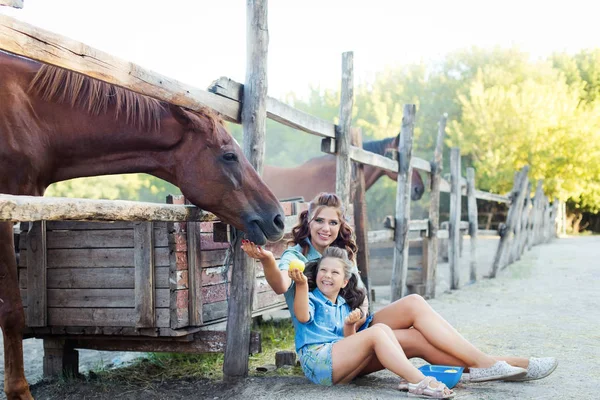 Una pequeña niña sonriente con madre con el pelo rizado vestida con vaqueros sentada en el suelo cerca de la cerca del caballo en el establo — Foto de Stock