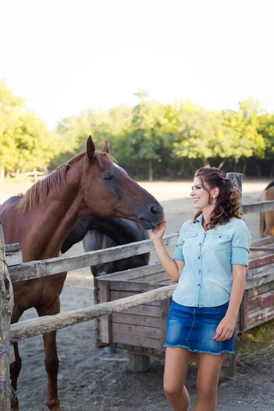 Una joven sonriente con el pelo rizado vestida con jeans en el establo con caballos — Foto de Stock