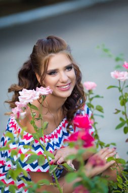 Gülümseyen kadın kahverengi kıvırcık saçlı mavi kırmızı pembe elbiseli zikzak baskı ile gül bahar yaz bahçede giyinmiş