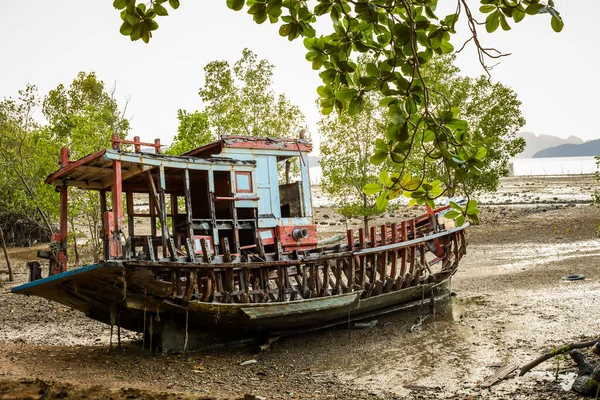 you can see an old colorful boat that is completely broken and lying on dry ground because there is low tide around it leaves and trees can be seen