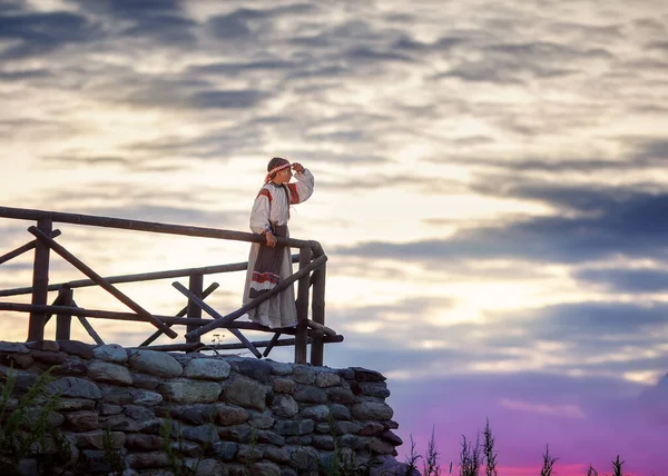 Beautiful girl in a traditional Russian dress on the background of the sunset. A young girl looks into the distance standing on the fence. Russian landscape.