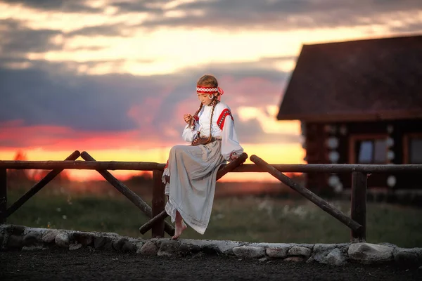 A beautiful girl in a traditional Russian dress is sitting on a fence against the sunset. The young girl thought about something. Traditional Russian costume.