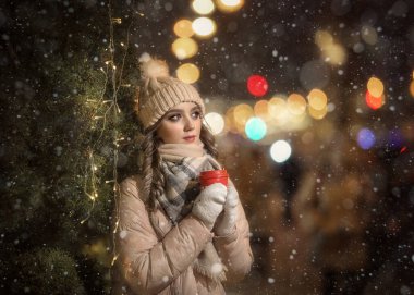 Eldivenli bir kız ve Noel ağacının yanında bir şapka. Bej ceketli bir kızın kış portresi. Elinde kırmızı kahve fincanı olan bir bayanın Noel portresi..