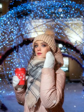 Noel çelenklerinden Bokeh 'in arka planındaki genç bayan. Güzel bir kızın Noel portresi. Bir kış gecesinde elinde bir fincan kahveyle bir kızın portresi..