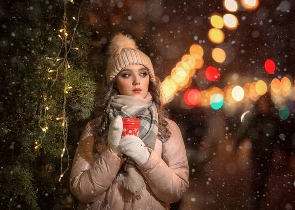 Eldivenli ve şapkalı bir kız kırmızı kahve bardağını Noel ağacının yanında tutuyor. Bej ceketli bir kızın kış portresi. Noel Portresi.
