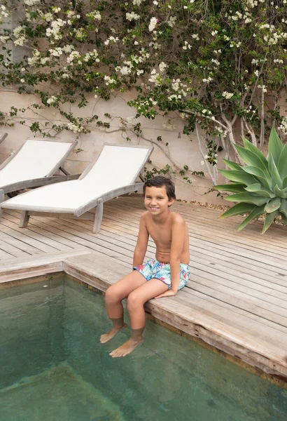 Shirtless boy in swimming shorts sitting at poolside looking at camera and enjoying his summer holidays