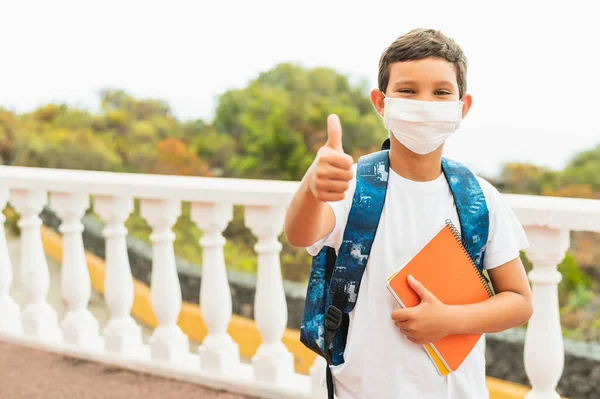 Kirlilik ya da virüs için koruyucu yüz maskesi takan okul çocuğunun portresi ve sabah okul otobüsünü beklerken başparmağını kaldırması. Coronavirus ve okul konsepti.