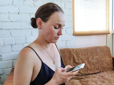 Kadın kanepede oturuyor, telefonu kullanıyor ve düşünüyor.