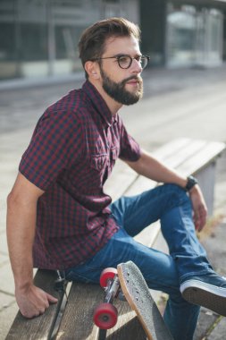 Sakallı adam bankta kaykayla oturuyor. 