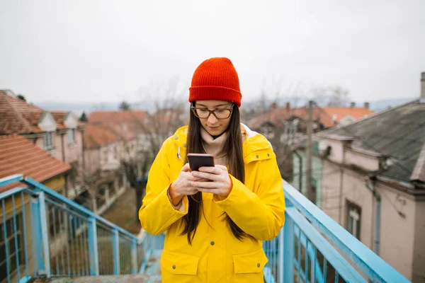 Millennial girl in raincoat chatting online on mobile phone. Millennial female texting message on smartphone