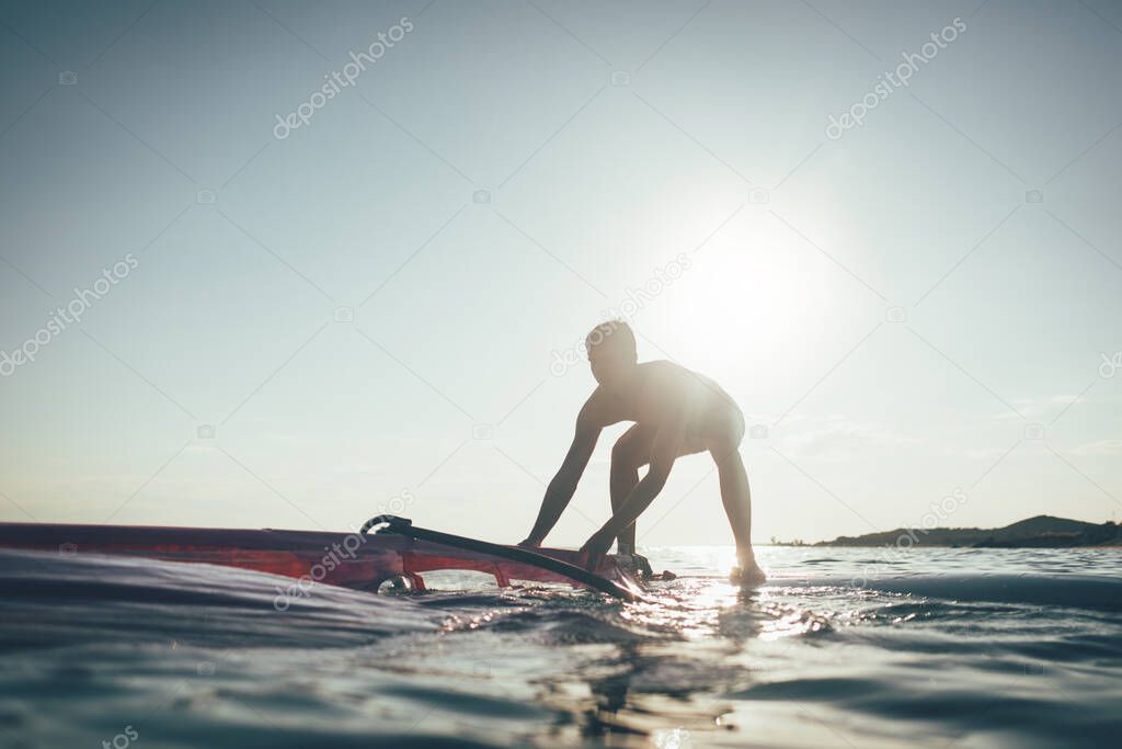 Young man uplift windsurf board sail. Surfer balancing on wind surf board on sunset sea. Windsurfing, summer, surfing, lifestyle