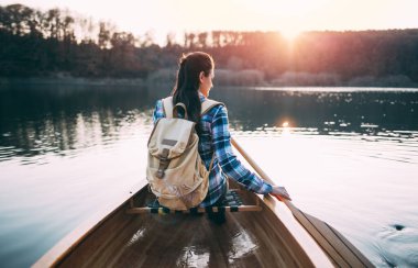 Sırt çantalı bir kızın günbatımı gölünde kanoda kürek çekişini. Açık havada iyi eğlenceler. Kano, kürek, spor