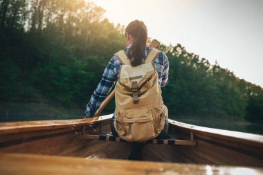 Sırt çantalı kız günbatımı gölünde tahta kanoda kürek çekiyor. Günbatımında sandalda kürek çeken gezgin kadının arka görüntüsü.