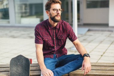 Sakallı adam bankta kaykayla oturuyor. 