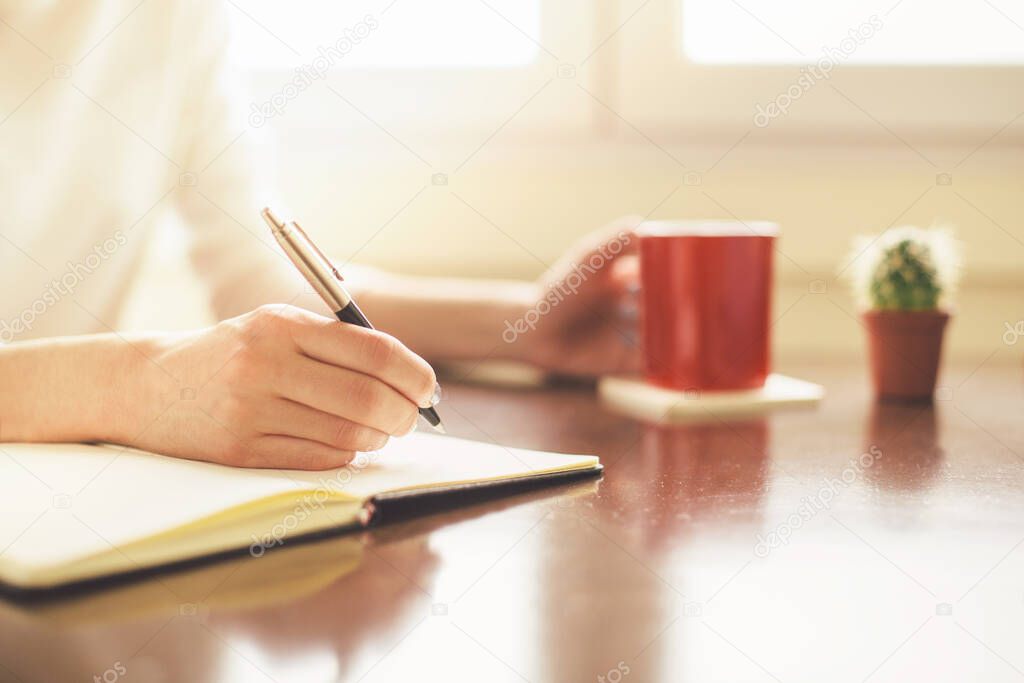 Cropped shot of woman writing and holding coffee cup