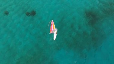 Rüzgâr sörfü tahtasında turkuaz denizde süzülen adamın hava görüntüsü. Rüzgarı yakalayan rüzgar sörfçüsünün görüntüsü.  