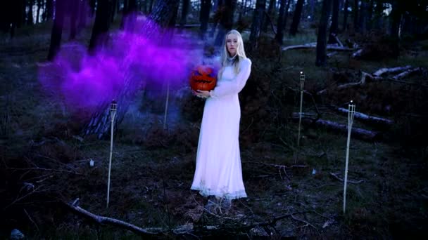 Hermosa rubia joven en un vestido blanco sosteniendo una calabaza con humo púrpura — Vídeos de Stock